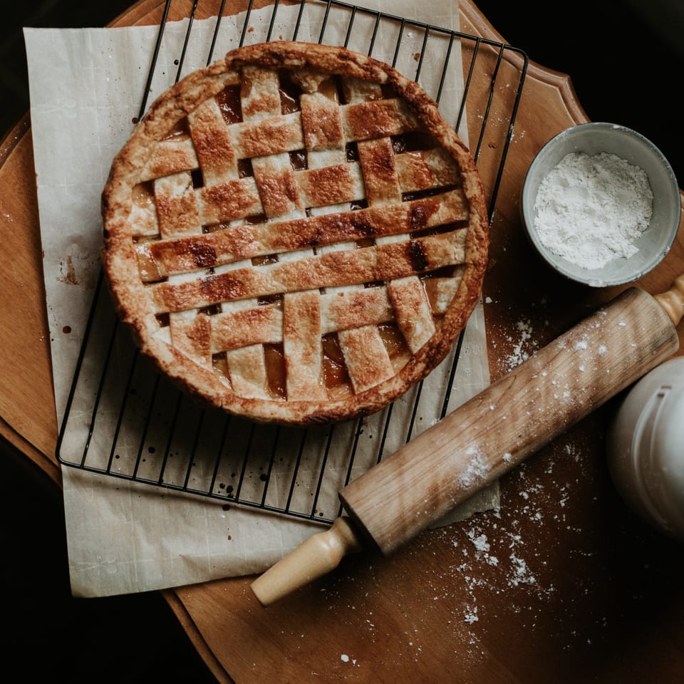 Freshly baked lattice-top pie on a cooling rack with a rolling pin and flour.