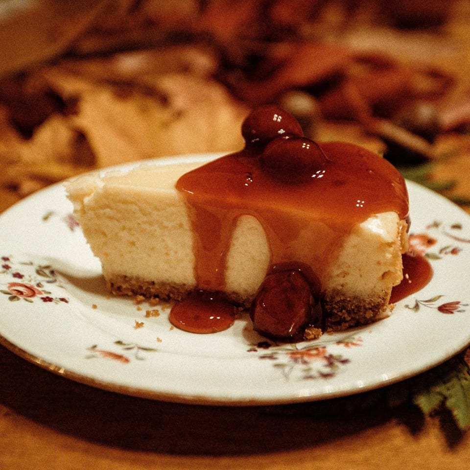 A slice of cheesecake with cherry topping and sauce on a white floral plate.