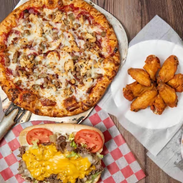 A pizza, a plate of chicken wings, and a cheesesteak sandwich on a wooden table.